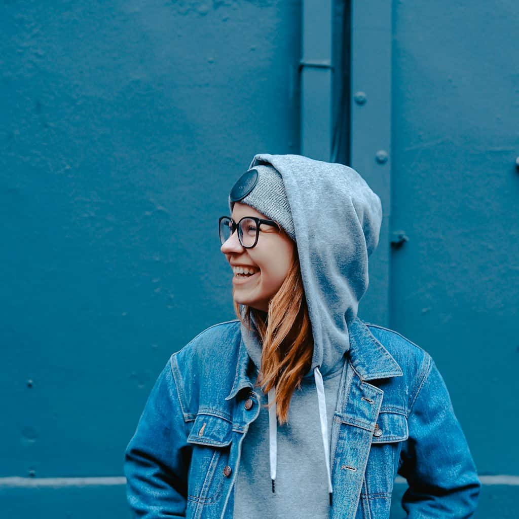 Smiling young woman in hoodie and glasses against blue wall.