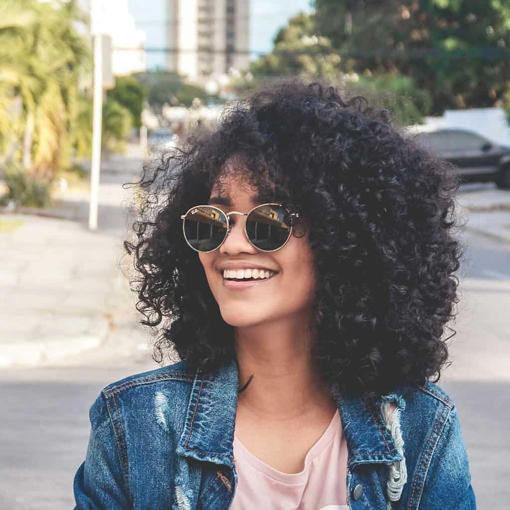 Woman with curly hair, sunglasses, and denim jacket smiling outdoors.