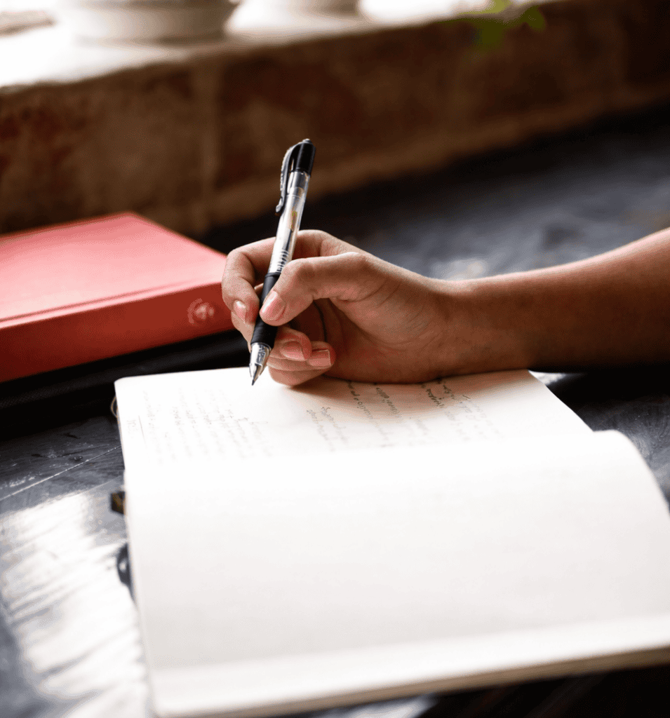 Person writing in a notebook with a pen on a wooden table.