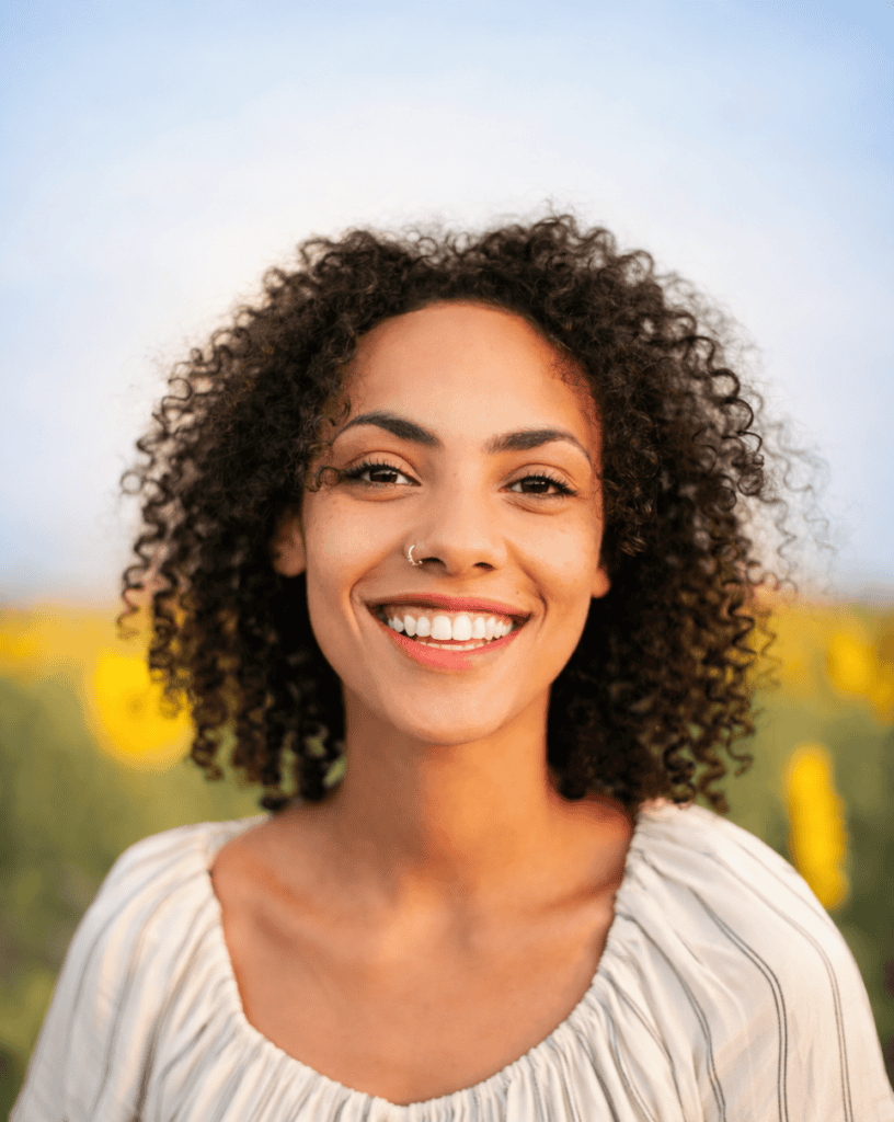 Smiling woman with curly hair in a sunflower field under a blue sky.