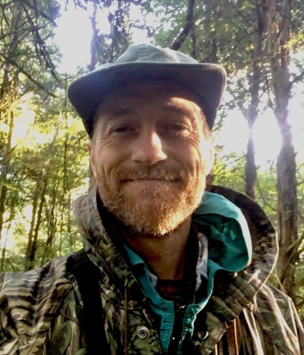 Smiling man in outdoor forest setting with hat and backpack.