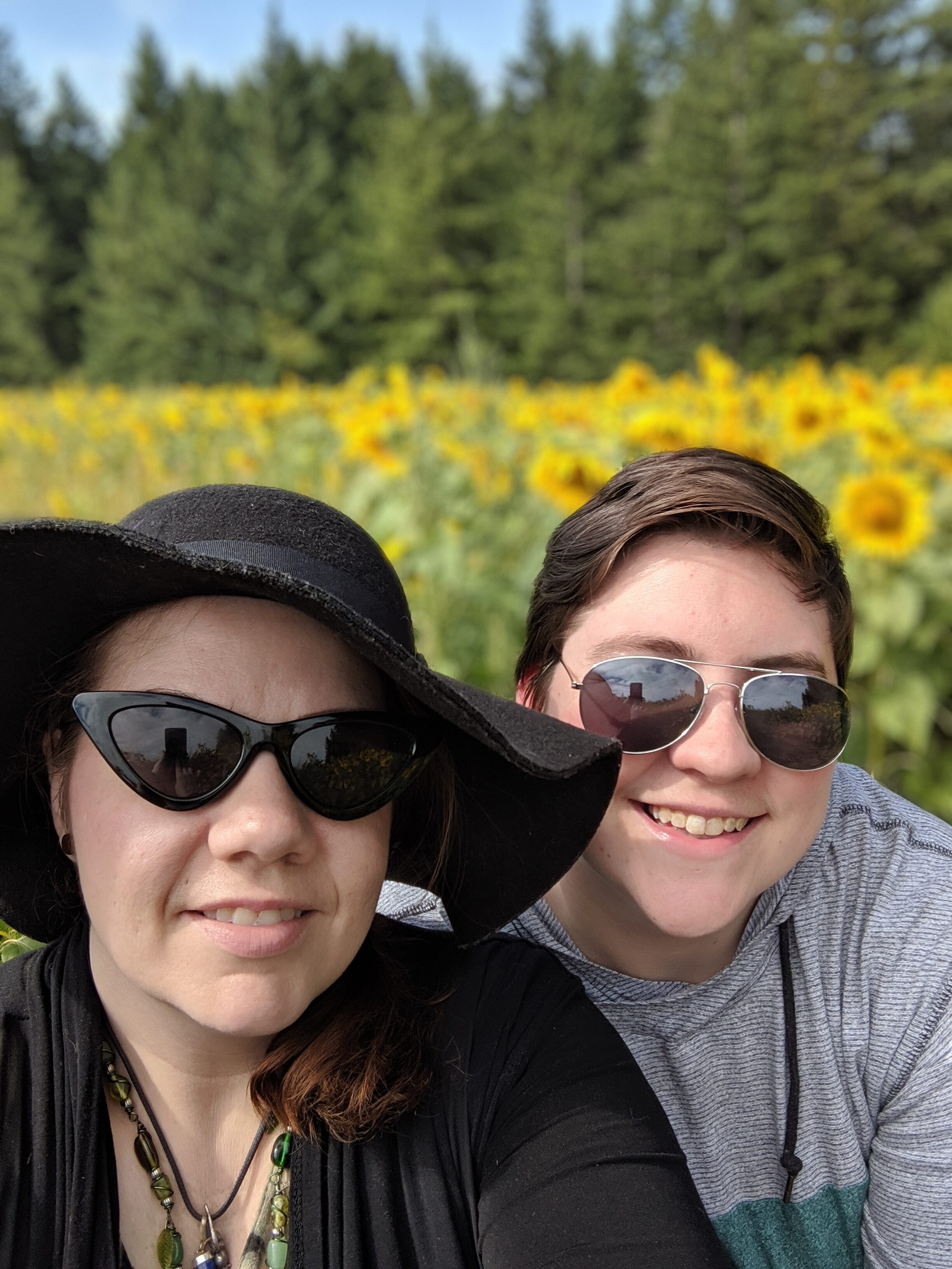 Smiling woman and young man in sunglasses outdoors with sunflowers in background.