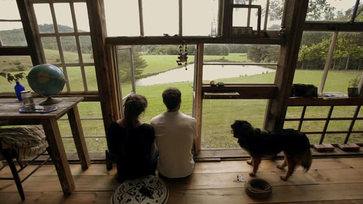 Couple sitting inside a rustic cabin looking at a pond and green landscape.