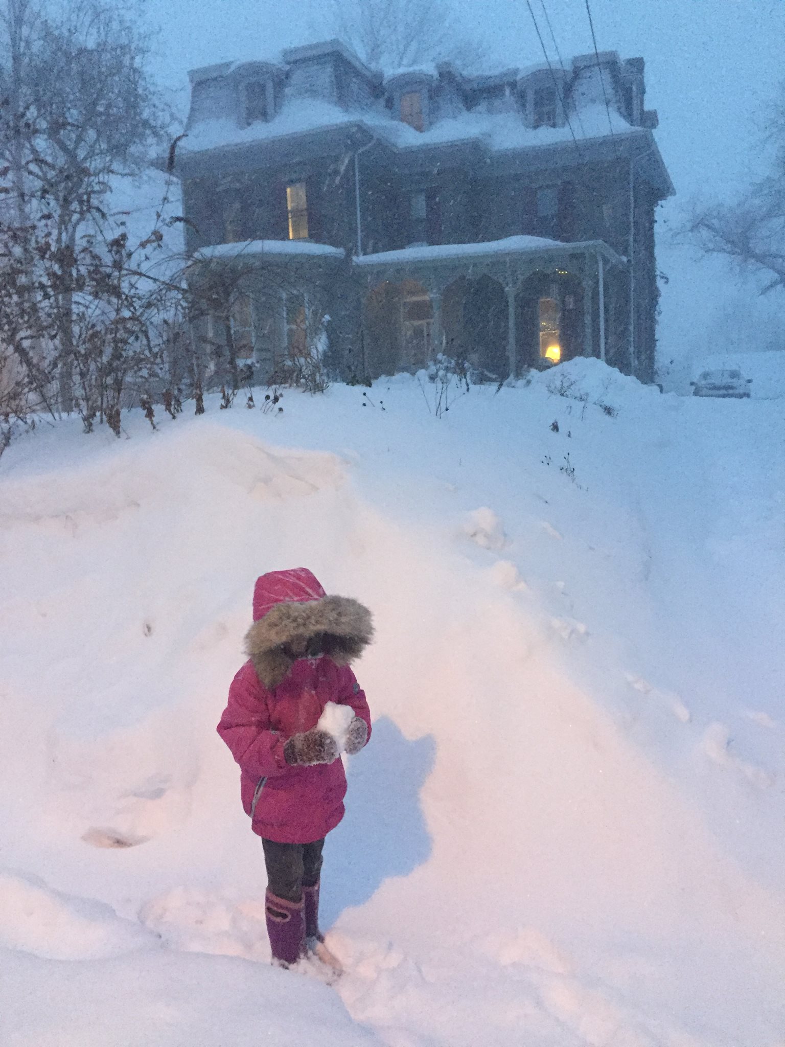 Child in pink winter coat standing in snow in front of a large house during winter.