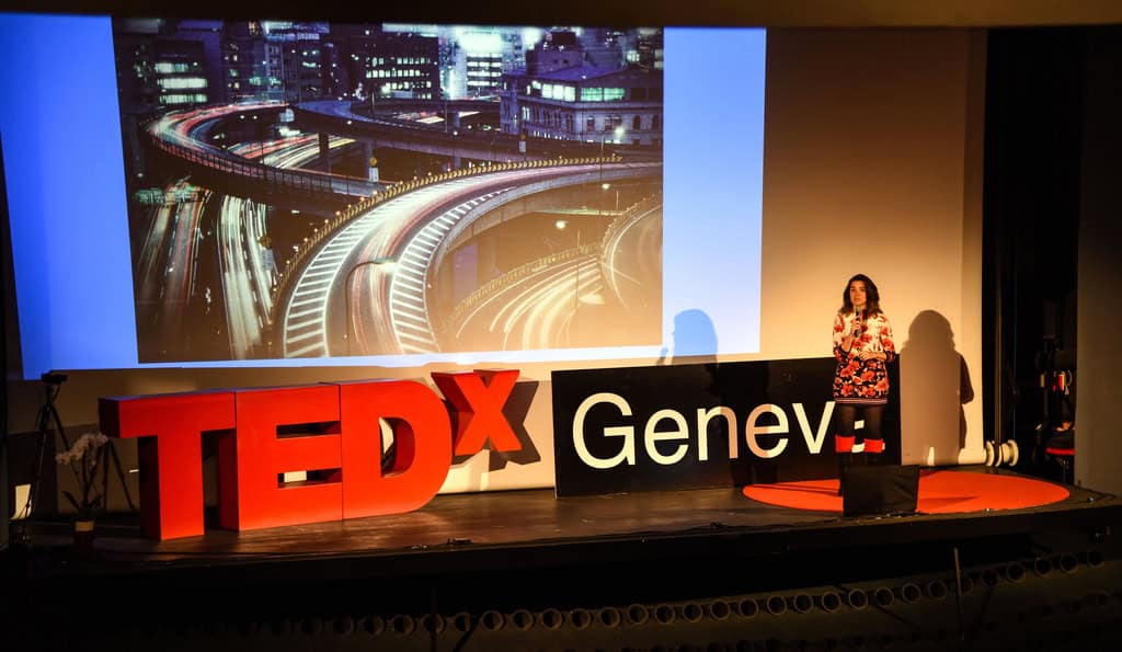 Speaker presenting at TEDxGeneva event with large screen and TEDx sign.