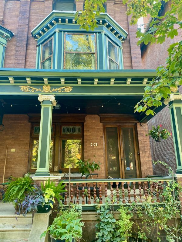 Historic Victorian house with green and gold accents, lush garden, and porch.