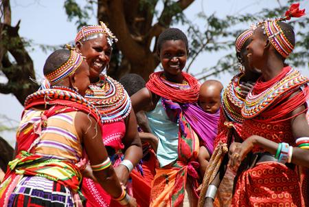 Women and children in traditional Maasai attire smiling and talking outdoors.