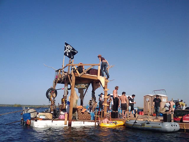 People gather on a floating wooden structure with a pirate flag on a sunny day.