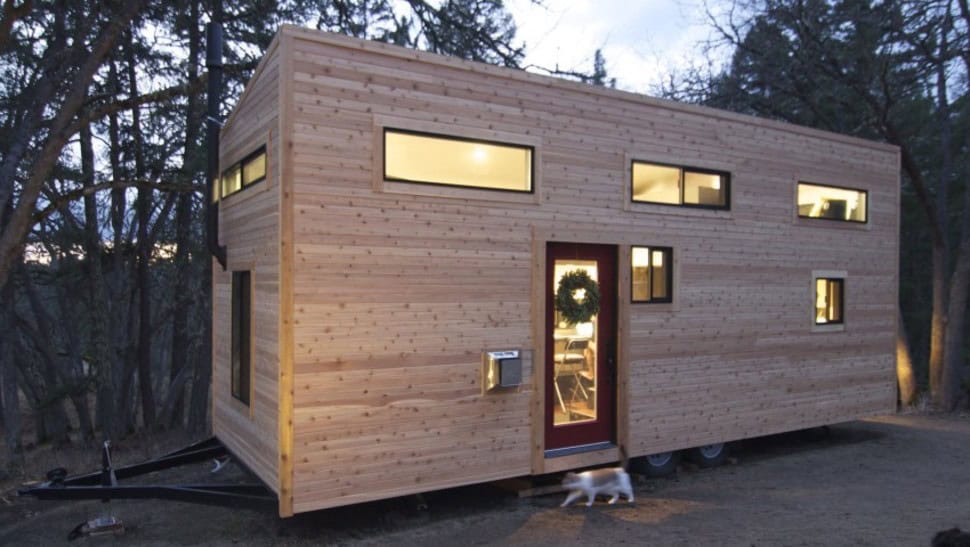 Modern tiny home with wooden exterior and multiple windows in a wooded area.