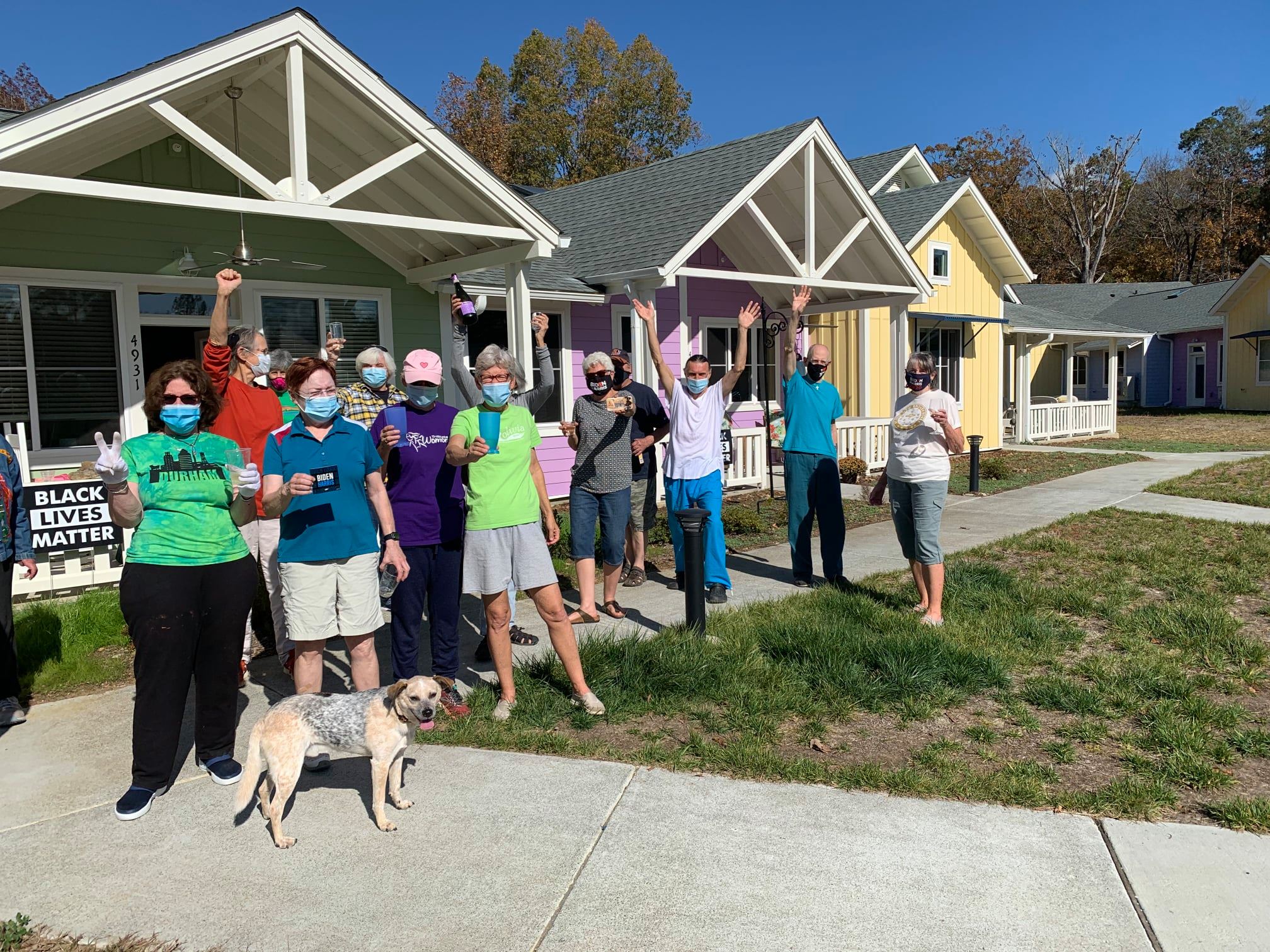 Group of diverse people with masks standing in front of colorful community houses.