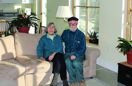 Elderly couple sitting on a beige sofa in a bright living room with plants.