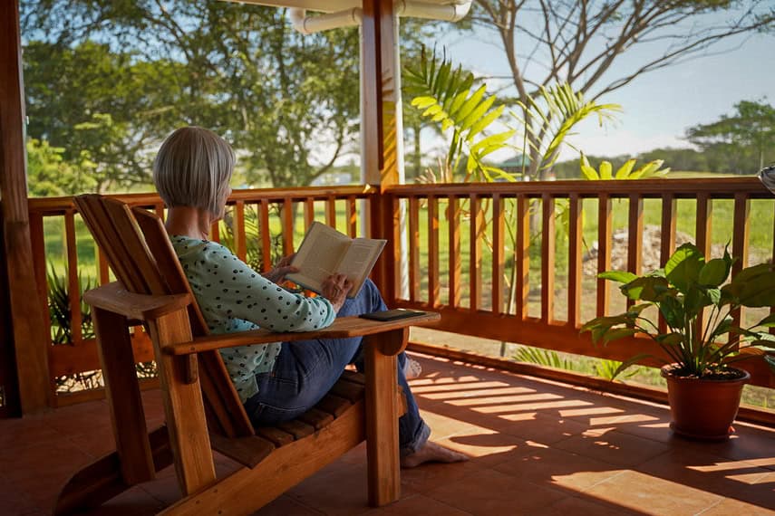 Woman reading a book on a wooden porch overlooking lush greenery.