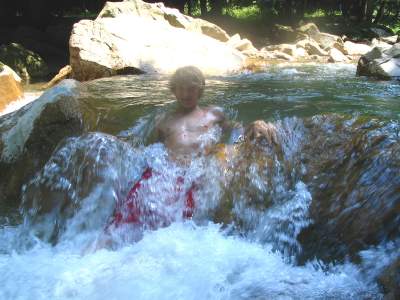 Young boy swimming in a natural river with rocks and trees in the background.