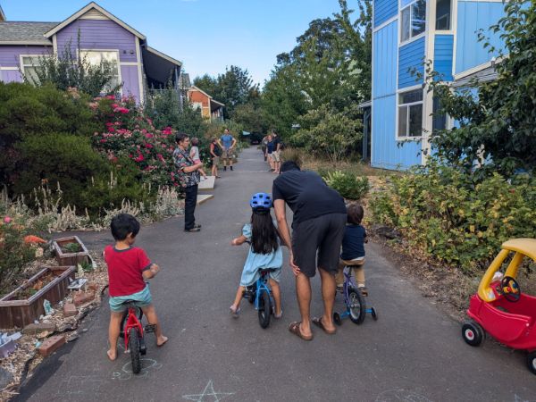 Community members and children riding bikes in a colorful eco-village setting.