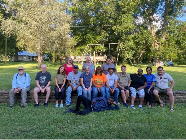 Group of diverse people sitting on a grassy field with trees and playground in background.