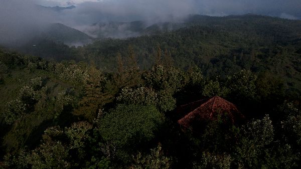 Lush green forested mountains with a small house and misty clouds in the background.