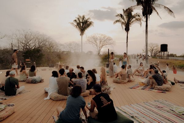 Group of people gathered outdoors on a wooden deck with palm trees in the background.