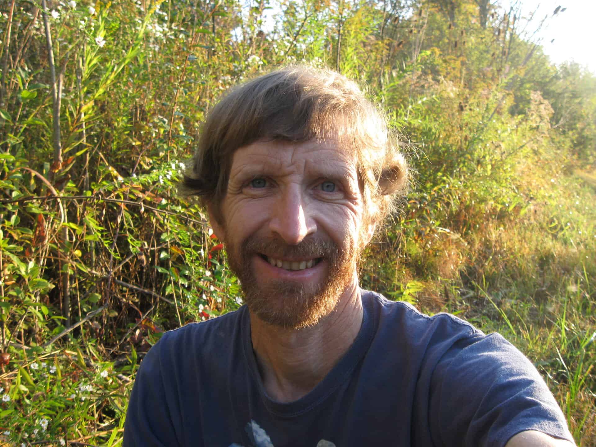 Smiling man outdoors in a natural setting with greenery and sunlight.