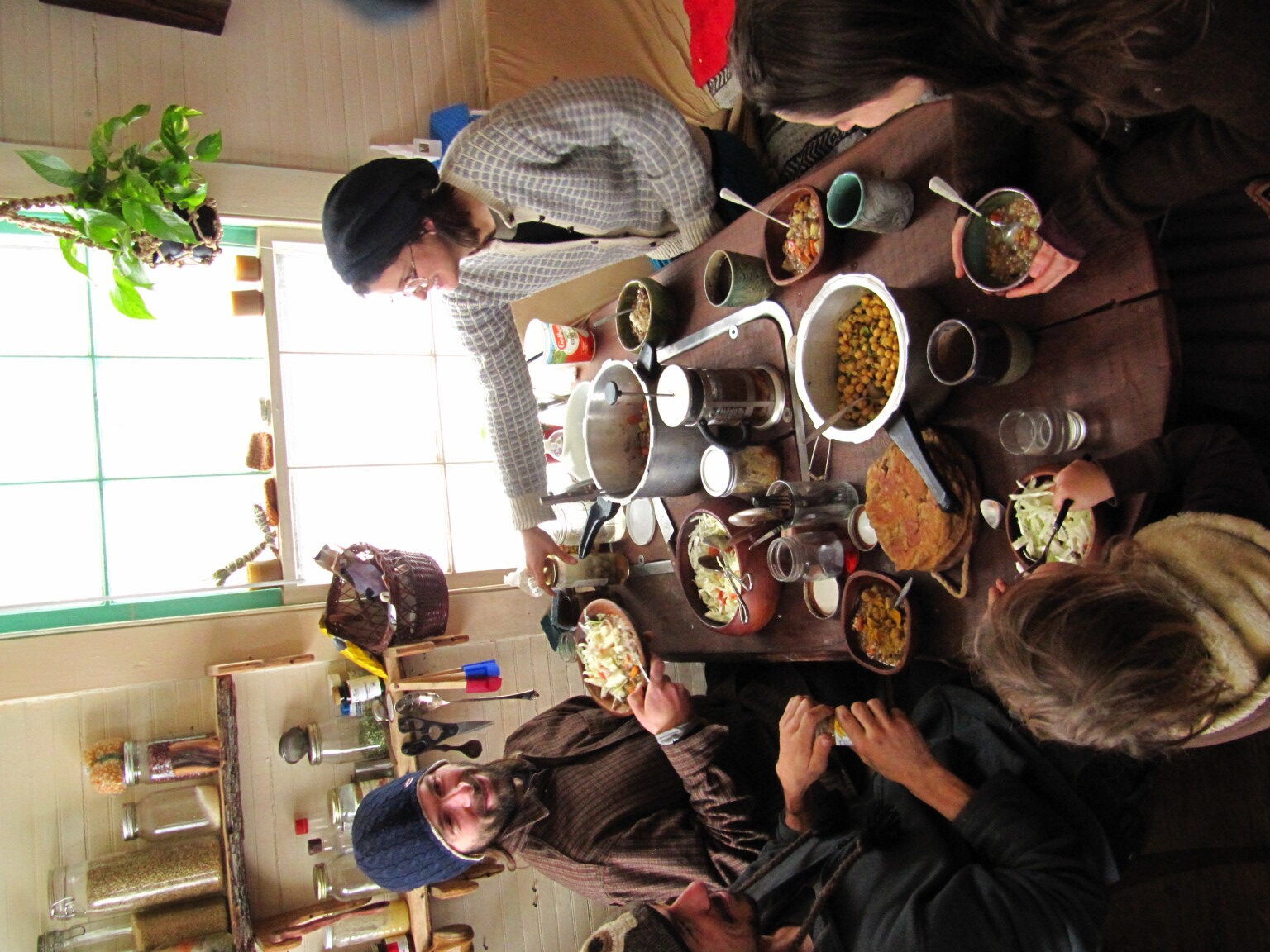 People enjoying a communal meal at a long wooden table in a cozy, well-lit kitchen.