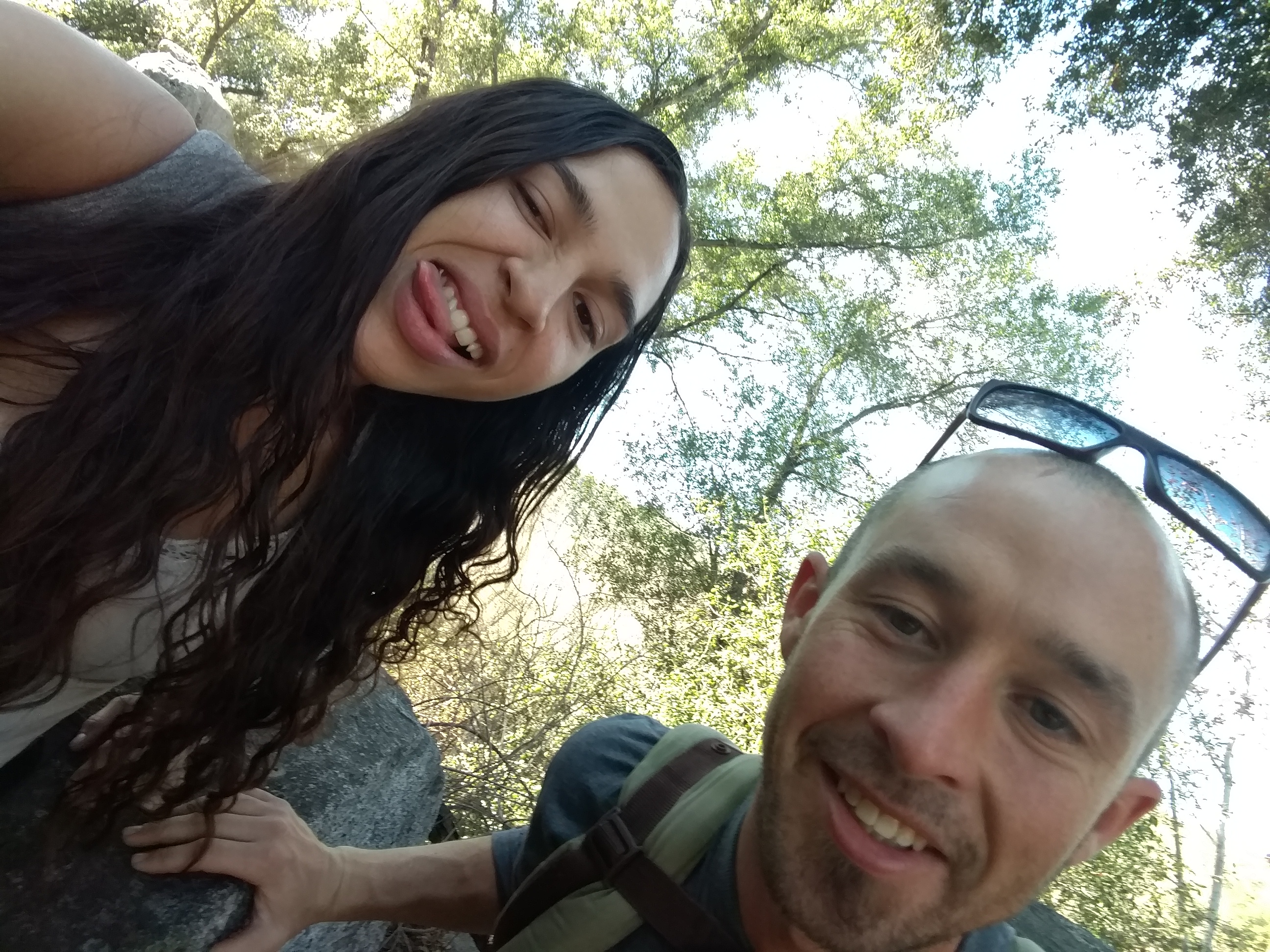 Smiling woman and man outdoors in a forest setting with sunlight filtering through trees.