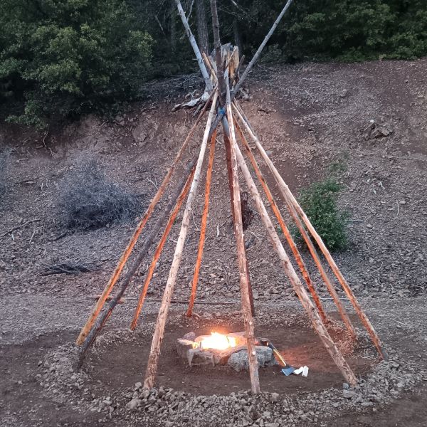 Campfire inside a teepee-style wooden structure in a natural setting.