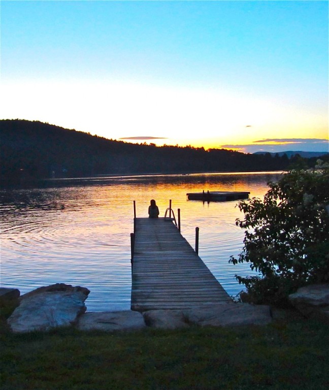 Serene lakeside scene with a wooden dock at sunset and a person sitting at the end.