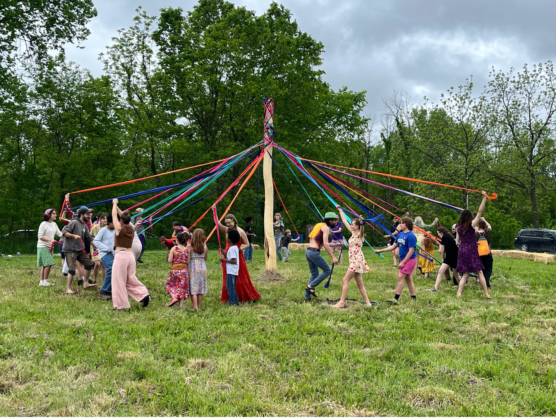 Children and adults dancing around a colorful maypole in a grassy field.