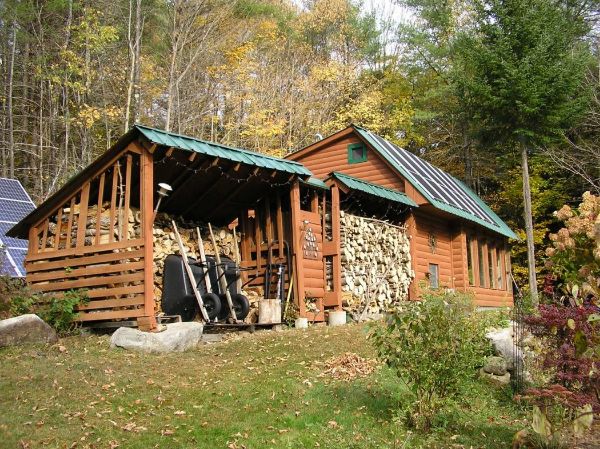 Rustic wooden cabin with solar panels and firewood storage in a forest setting.