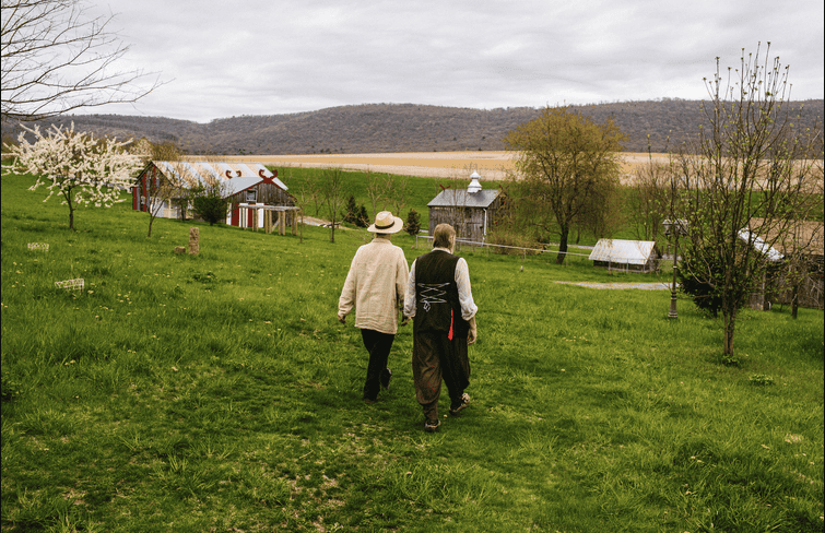 People walking through a rural community with farm buildings and trees under cloudy sky.