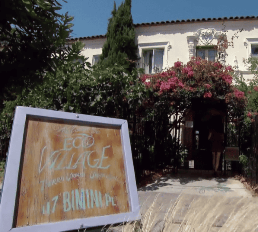 Eco Village sign in front of a white house with pink flowering bushes.