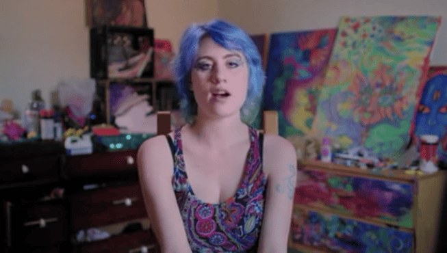 Young woman with blue hair speaking in a room with colorful paintings.