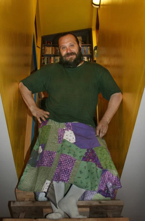 Man with a beard wearing a green shirt and colorful patchwork skirt in a narrow hallway.