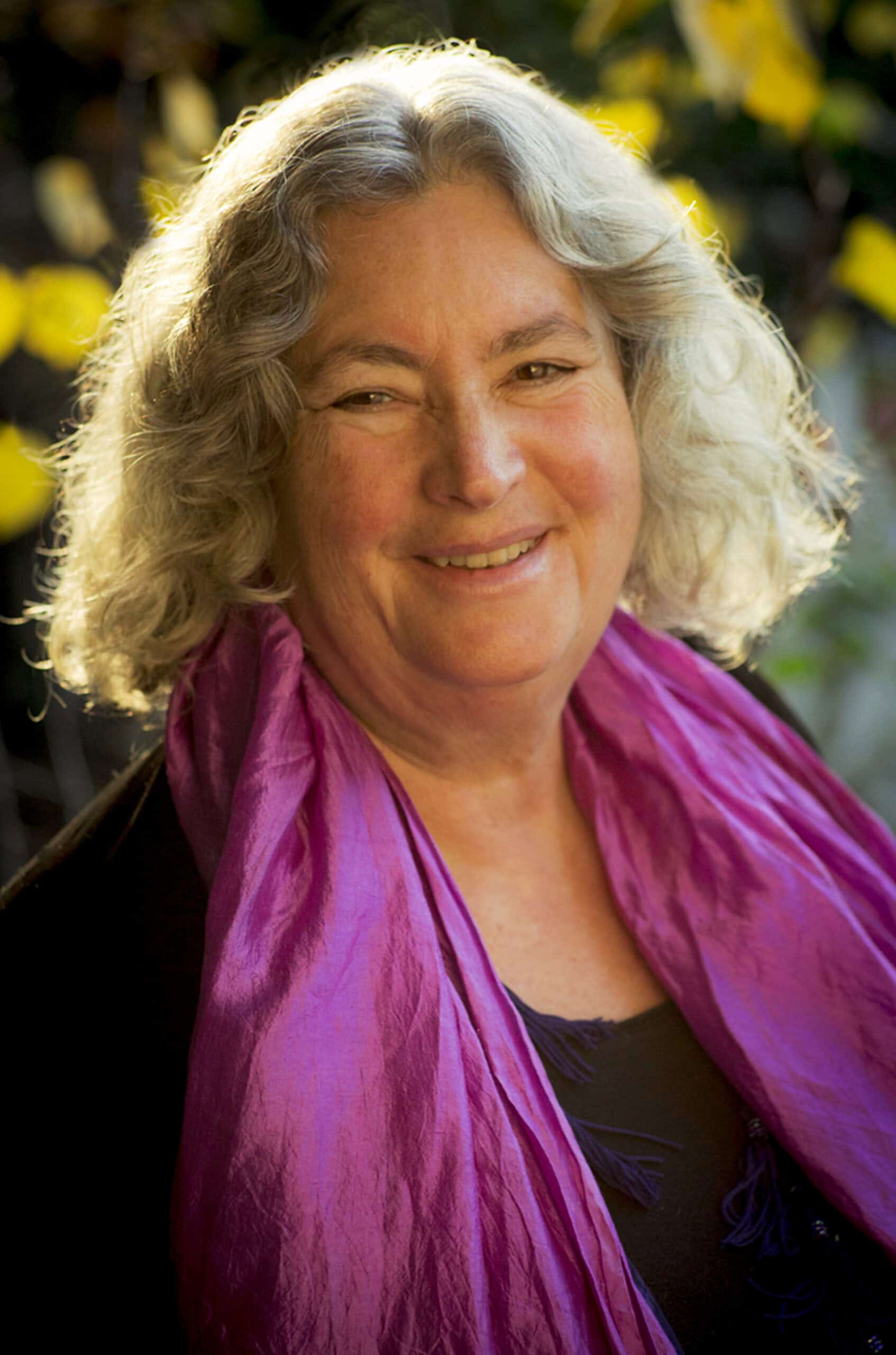 Smiling woman with curly gray hair wearing a purple scarf outdoors.