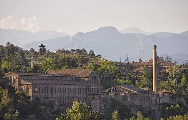 Historic industrial building surrounded by greenery with mountains in the background.