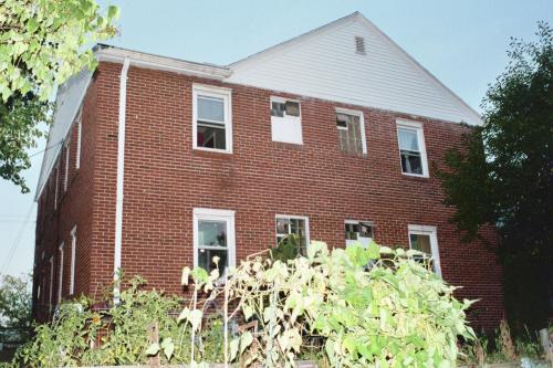 Brick building with white upper section and multiple windows, surrounded by greenery.