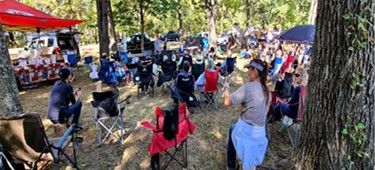 Community gathering at outdoor event in a wooded area with chairs and tables.
