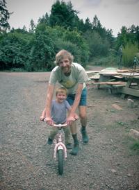 Adult helping a child ride a pink bicycle outdoors in a natural setting.
