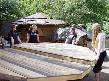Group of people building a large wooden structure outdoors in a green setting.