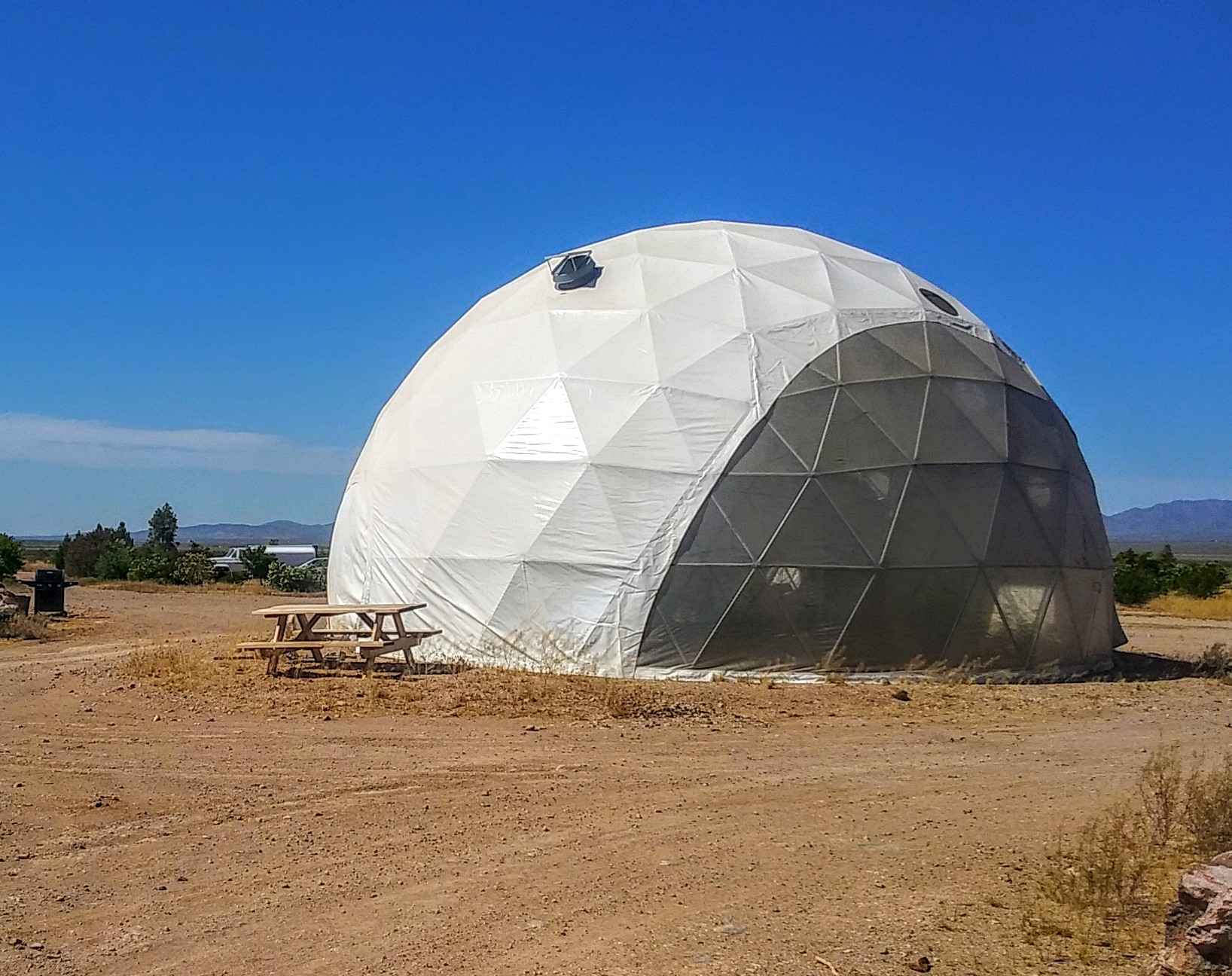 Geodesic dome structure on a desert landscape with picnic table and mountains in background.