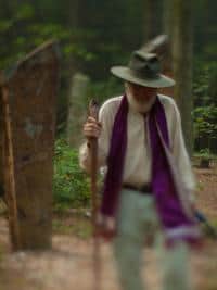 Person in forest wearing hat and scarf, holding a walking stick.