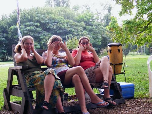 Three women sitting on a wooden swing in a park, making hand gestures.