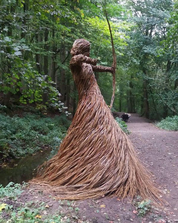 Sculpture of a woman with a bow in a forest setting, made from natural materials.