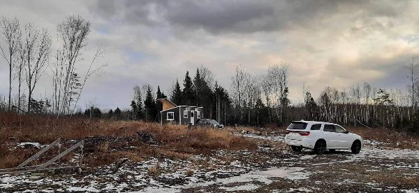 Rural landscape with a small house and a white SUV parked outside under cloudy sky.