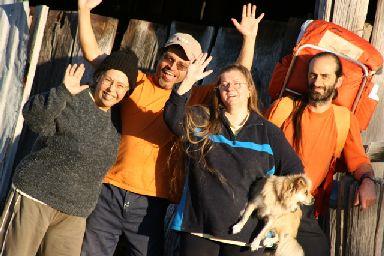 Group of four diverse people smiling and waving outdoors with a dog and backpack.