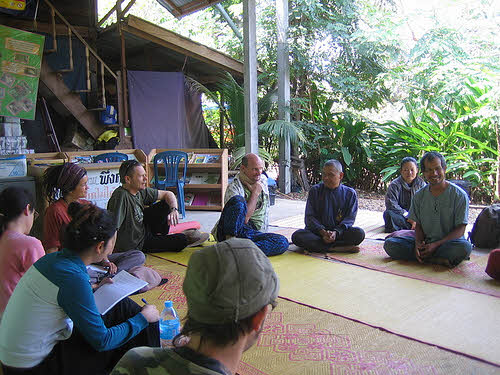 Group of people sitting on mats in a lush outdoor setting for a community discussion.