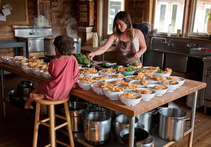 Woman serving food to a child at a community kitchen table with many dishes.