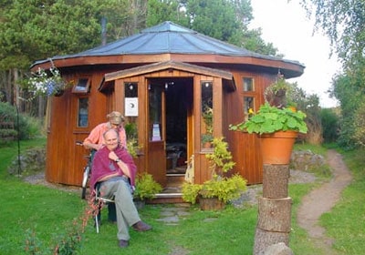 Wooden eco-friendly tiny house with garden and elderly woman sitting outside.