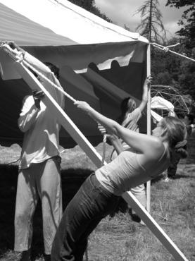 People setting up a large outdoor tent in a forested area.