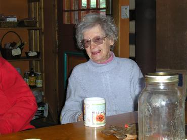 Elderly woman with glasses smiling at a table in a cozy home setting.