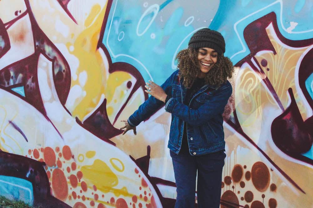 Smiling woman in denim jacket and beanie in front of colorful graffiti wall.